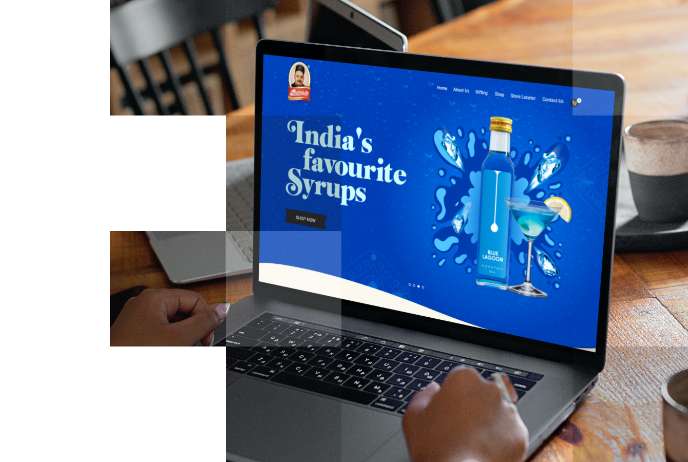 A silver laptop computer displaying a website with the text "India's favourite Syrups. Shop Now Lagoona" on the screen. A bottle of syrup is on top of the laptop screen, obscuring part of the text. The laptop is on a wooden table.